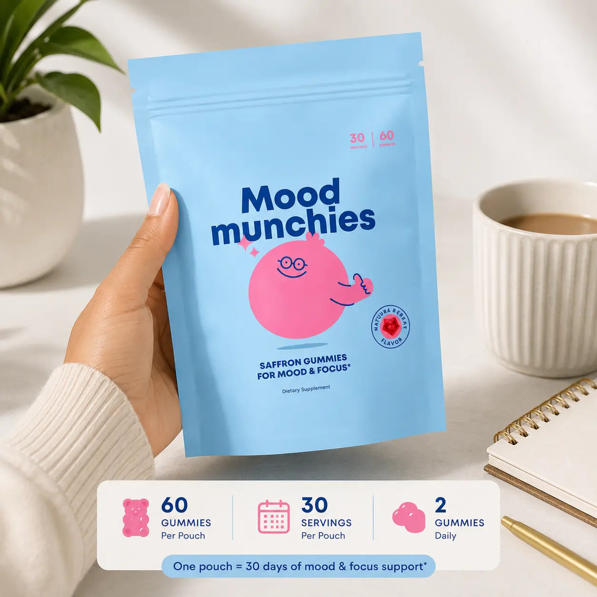 A hand holds a light blue stand-up pouch labeled 'Mood munchies' with a pink cartoon character. The scene is styled on a white surface with a plant, a cup of coffee, a notebook, and a pen in the background.